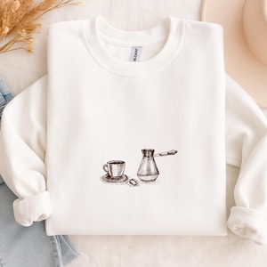 Puede incluir: Sudadera blanca con un dibujo detallado a pluma y tinta de una taza de café, un platillo, granos de café y una cafetera tradicional. El diseño está centrado en la parte delantera de la parte superior de cuello redondo y manga larga.