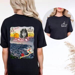 Puede incluir: Una camiseta negra con un gr&aacute;fico en la espalda y un logotipo peque&ntilde;o en la parte delantera. El gr&aacute;fico trasero muestra un retrato de Fairuz, el texto "alo beirut" en ingl&eacute;s y &aacute;rabe, notas musicales, un atardecer y un coraz&oacute;n con la bandera.