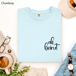 Puede incluir: Una camiseta de cuello redondo azul claro con el texto "alo beirut" impreso en letra cursiva negra en la parte inferior derecha. La camiseta cuenta con una etiqueta de la marca Comfort Colors en el cuello interior y es de color chambray.