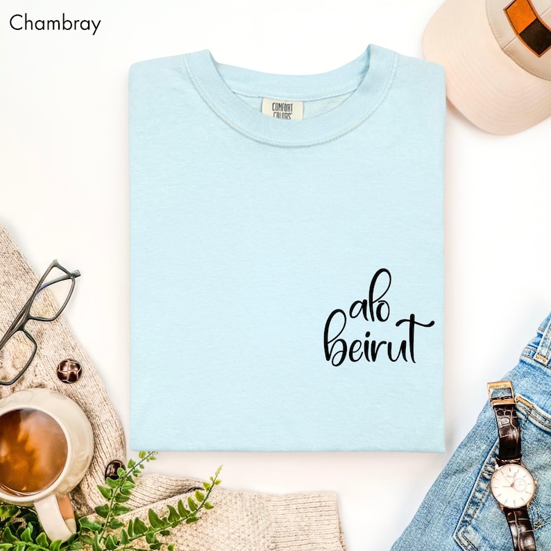 Puede incluir: Una camiseta de cuello redondo azul claro con el texto "alo beirut" impreso en letra cursiva negra en la parte inferior derecha. La camiseta cuenta con una etiqueta de la marca Comfort Colors en el cuello interior y es de color chambray.
