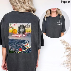 Puede incluir: Una camiseta gris carb&oacute;n con un gr&aacute;fico en la espalda y un logotipo peque&ntilde;o en el frente. El gr&aacute;fico trasero muestra el retrato de una mujer, un atardecer sobre una ciudad, notas musicales, el texto "alo beirut" en ingl&eacute;s y &aacute;rabe, y un coraz&oacute;n con la bandera libanesa.