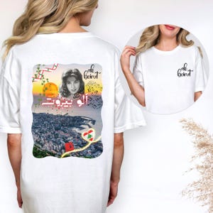 Puede incluir: Una camiseta blanca con un gr&aacute;fico en la espalda y un peque&ntilde;o logotipo en la parte delantera. El gr&aacute;fico de la espalda muestra el retrato de una mujer, un paisaje urbano de Beirut, notas musicales, un coraz&oacute;n con la bandera libanesa y el texto Alo Beirut.