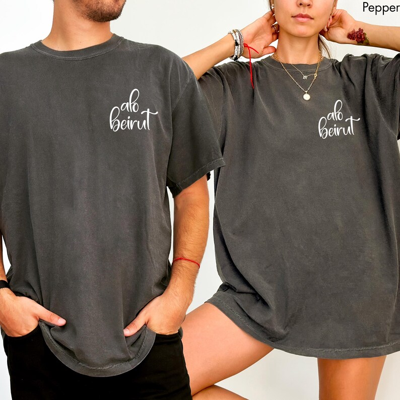 Puede incluir: Una camiseta de manga corta gris carb&oacute;n con el texto cursivo blanco "alo beirut" impreso en la parte superior izquierda del pecho. La prenda tiene cuello redondo, un corte holgado y una textura de tela desgastada de estilo vintage.