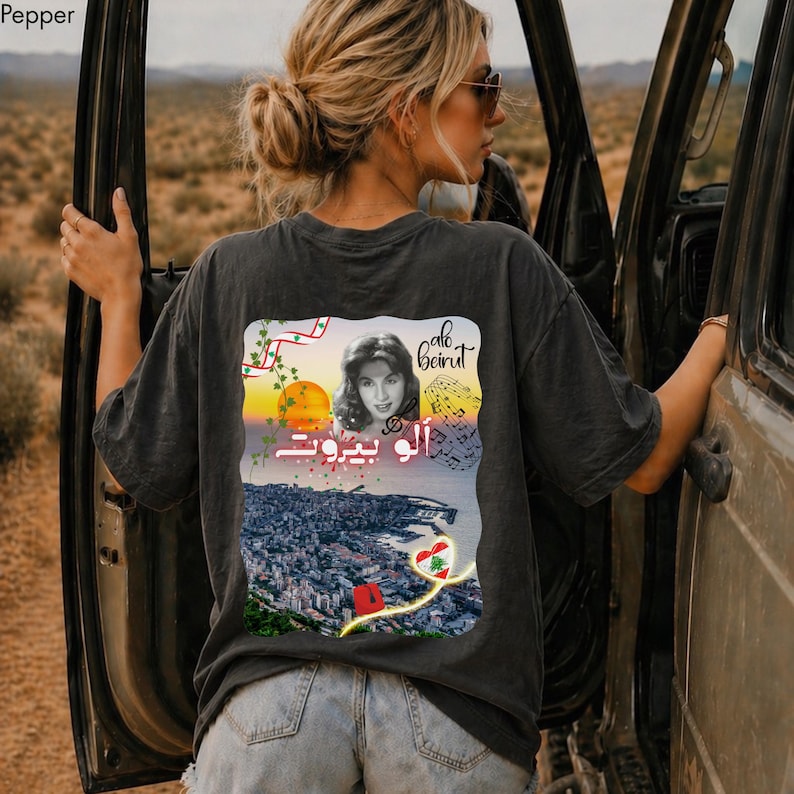 Puede incluir: Una camiseta gris carb&oacute;n de manga corta con un gr&aacute;fico rectangular en la espalda. El estampado muestra el retrato de una mujer, un atardecer sobre una ciudad costera, notas musicales, el texto "alo Beirut" y un coraz&oacute;n con un cedro.