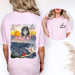 Puede incluir: Una camiseta rosa claro con un gr&aacute;fico en la parte posterior y un logotipo m&aacute;s peque&ntilde;o en el frente. El gr&aacute;fico posterior muestra el retrato de una mujer, notas musicales, un atardecer sobre una ciudad costera y el texto "alo beirut" en ingl&eacute;s y &aacute;rabe.