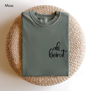 Puede incluir: Una camiseta de color verde musgo con cuello redondo, cuello acanalado y el texto cursivo negro "alo beirut" impreso en la parte inferior derecha. La camiseta tiene una etiqueta de la marca Comfort Colors en el interior del cuello.