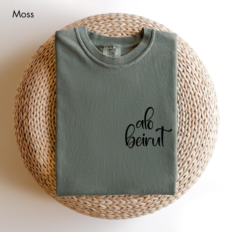 Puede incluir: Una camiseta de color verde musgo con cuello redondo, cuello acanalado y el texto cursivo negro "alo beirut" impreso en la parte inferior derecha. La camiseta tiene una etiqueta de la marca Comfort Colors en el interior del cuello.