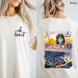 Puede incluir: Una camiseta color marfil con el texto "alo beirut" en letra cursiva negra en la parte delantera. La parte trasera muestra un gr&aacute;fico con el retrato de una mujer, notas musicales, un atardecer sobre una ciudad costera, la bandera libanesa y el texto.