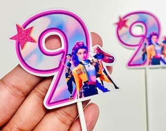May include: Two number nine cake toppers. Each topper features a pink number nine with a star and a character illustration. The characters are wearing yellow and blue jackets. One topper is held by a hand.