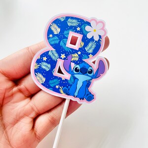 Puede incluir: Adorno para tarta con el n&uacute;mero ocho, decorado con un estampado floral azul y un personaje de dibujos animados. El n&uacute;mero est&aacute; delineado en rosa y tiene una flor blanca. El personaje es azul.