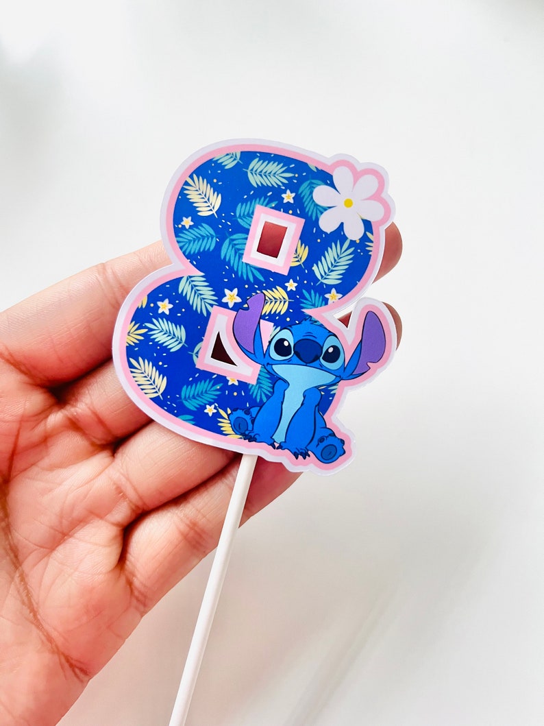Puede incluir: Adorno para tarta con el n&uacute;mero ocho, decorado con un estampado floral azul y un personaje de dibujos animados. El n&uacute;mero est&aacute; delineado en rosa y tiene una flor blanca. El personaje es azul.