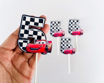 May include: Four red race car cake toppers with a black and white checkered number 5. The toppers are on white sticks.