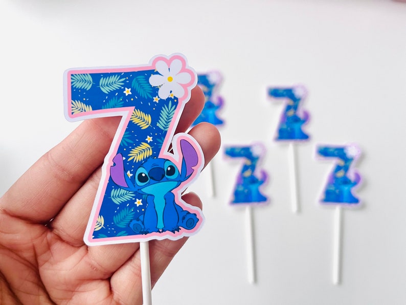 Puede incluir: Adorno para tarta con el n&uacute;mero siete en azul y rosa, con el personaje Stitch de Lilo & Stitch. El n&uacute;mero est&aacute; decorado con un estampado de hojas tropicales y una flor blanca. Se ven adornos adicionales en el fondo.