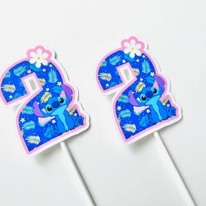 Puede incluir: Dos adornos para tartas con forma del n&uacute;mero 2, con el personaje Stitch de Lilo & Stitch. El n&uacute;mero es azul con un estampado de hojas y un borde rosa. Stitch est&aacute; en el centro y una flor blanca en la parte superior. En palitos blancos.