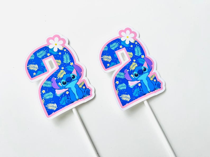 Puede incluir: Dos adornos para tartas con forma del n&uacute;mero 2, con el personaje Stitch de Lilo & Stitch. El n&uacute;mero es azul con un estampado de hojas y un borde rosa. Stitch est&aacute; en el centro y una flor blanca en la parte superior. En palitos blancos.