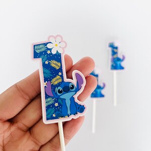 Puede incluir: Un adorno para tarta azul con el n&uacute;mero uno y el personaje Stitch de Lilo & Stitch. El n&uacute;mero est&aacute; decorado con un estampado de hojas tropicales y una flor blanca. Se ven adornos adicionales en el fondo.
