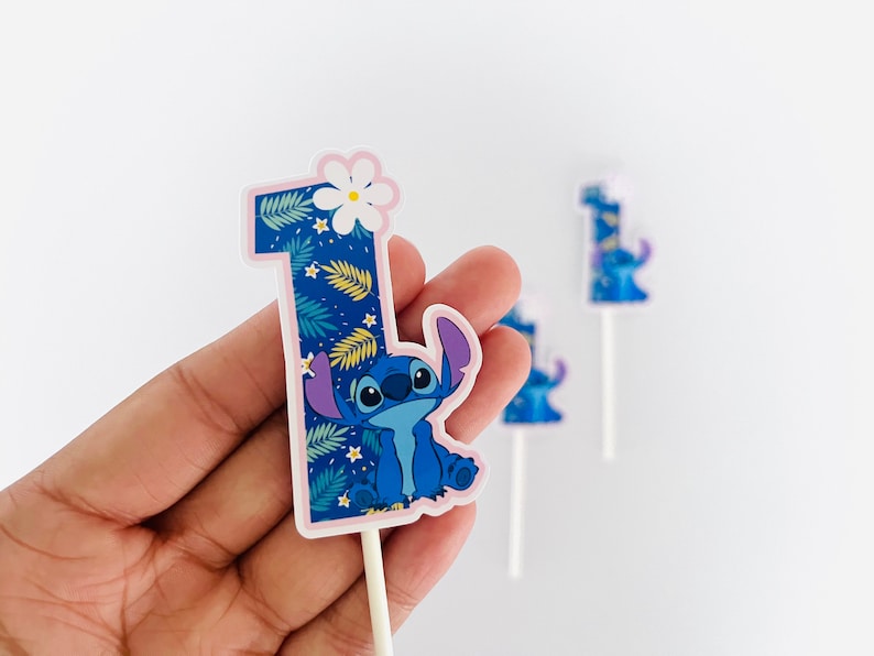 Puede incluir: Un adorno para tarta azul con el n&uacute;mero uno y el personaje Stitch de Lilo & Stitch. El n&uacute;mero est&aacute; decorado con un estampado de hojas tropicales y una flor blanca. Se ven adornos adicionales en el fondo.