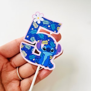 Puede incluir: Adorno para tarta con el n&uacute;mero 5 y Stitch de Lilo & Stitch. El n&uacute;mero es azul con un estampado de hojas tropicales y un borde rosa. Una flor blanca est&aacute; en la parte superior. Stitch est&aacute; en la parte inferior.