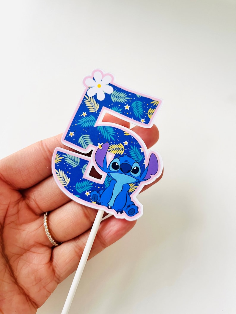 Puede incluir: Adorno para tarta con el n&uacute;mero 5 y Stitch de Lilo & Stitch. El n&uacute;mero es azul con un estampado de hojas tropicales y un borde rosa. Una flor blanca est&aacute; en la parte superior. Stitch est&aacute; en la parte inferior.