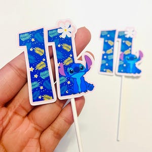 Puede incluir: Dos adornos para tartas azules y blancos con el n&uacute;mero 11 y un personaje de dibujos animados. Los n&uacute;meros tienen un patr&oacute;n de hojas tropicales y estrellas. Un adorno tiene el personaje Stitch. El otro adorno tiene una flor blanca.