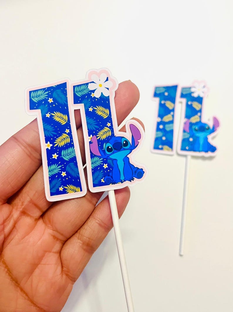 Puede incluir: Dos adornos para tartas azules y blancos con el n&uacute;mero 11 y un personaje de dibujos animados. Los n&uacute;meros tienen un patr&oacute;n de hojas tropicales y estrellas. Un adorno tiene el personaje Stitch. El otro adorno tiene una flor blanca.