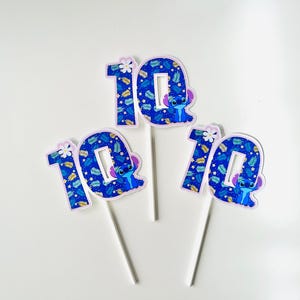 Puede incluir: Tres adornos para tartas con el n&uacute;mero 10, con un personaje de dibujos animados y un estampado de hojas azules. Cada n&uacute;mero est&aacute; sobre un palo blanco. Los adornos son azules, blancos y morados.