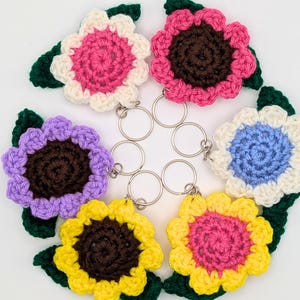 May include: A collection of six handmade crochet flower keychains in various colors: pink, purple, yellow, and blue. Each flower has a brown center, white or cream petals, and green leaves. The keychains are arranged in a circular pattern.