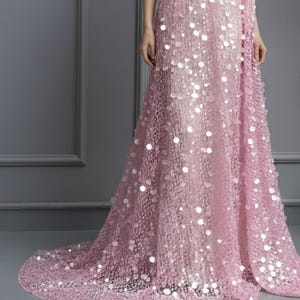 May include: A light pink, one-shoulder evening gown with a long train. The dress is covered in sequins, creating a shimmering effect. The model is wearing the dress in front of a gray wall.