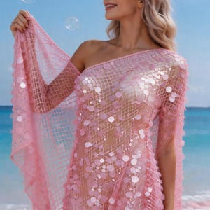 May include: A pink, one-shoulder dress with a net-like design and large, round sequins. The dress has a long sleeve and is worn on a beach with a blue sky and bubbles in the background. The dress is a light, airy garment.