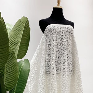 May include: White lace fabric with a delicate floral pattern. The fabric is draped over a mannequin and has a flowing, asymmetrical shape.