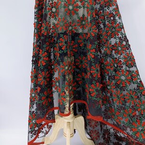May include: Red and green floral embroidered fabric on a black mesh background. The fabric is draped over a mannequin and has a red trim around the edges.