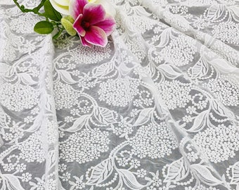 Pode incluir: Tecido de renda branca bordada com um delicado padrão floral. O tecido é transparente e tem uma textura macia e fluida.