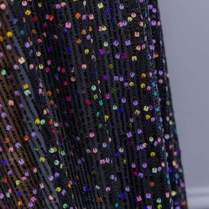 May include: Close-up of black fabric with colourful sequins. The sequins are in various shapes, including round and square, and are arranged in vertical rows. The fabric has a sheer quality, and the sequins reflect light, creating a shimmering effect.