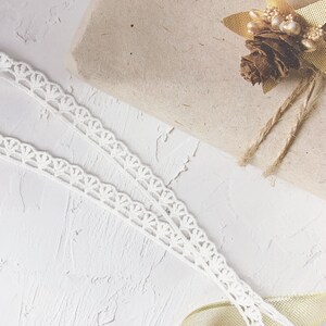 K&ouml;nnte beinhalten: Eine braune Geschenkbox aus Papier, die mit Schnur umwickelt und mit einer goldenen Tannenzapfen und Bl&auml;ttern verziert ist. Die Box liegt auf einer wei&szlig;en, strukturierten Oberfl&auml;che mit wei&szlig;er Spitzenborte.