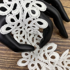 May include: Close-up of white lace trim with a detailed, swirling floral design. The lace is displayed on a black mannequin hand against a wooden background. The trim features a repeating pattern of interconnected loops and curves.