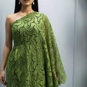 Può includere: Un lungo abito monospalla verde oliva con uno strascico fluente. L'abito è realizzato in un delicato tessuto di pizzo fantasia. La modella indossa orecchini pendenti e ha lunghi capelli scuri.