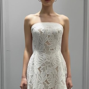 May include: A strapless white wedding gown with a floral lace overlay. The dress features a fitted bodice and a flowing skirt that extends into a small train. The model is standing in front of a gray wall.