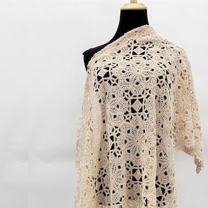 以下が含まれることがあります： 薄いベージュ色の繊細なクロシェのショール。レース模様で、わずかに非対称な形をしています。