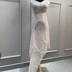 K&ouml;nnte beinhalten: Ein wei&szlig;er und rosa paillettenbesetzter Stoff mit einem High-Low-Saum. Der Stoff ist &uuml;ber eine Schaufensterpuppe drapiert.