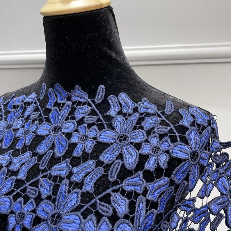 May include: A garment featuring intricate blue floral lace embroidery layered over a solid black fabric base. The lace pattern consists of detailed flower motifs with interconnected stems and leaves, covering the upper chest and shoulder area.