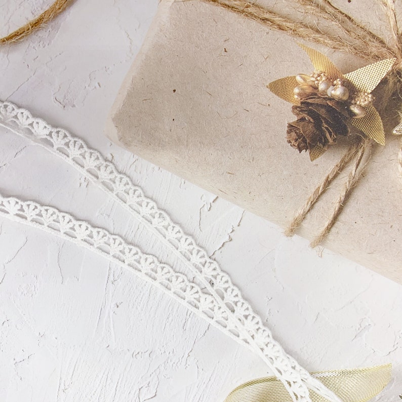 K&ouml;nnte beinhalten: Eine braune Geschenkbox aus Papier, die mit Schnur umwickelt und mit einer goldenen Tannenzapfen und Bl&auml;ttern verziert ist. Die Box liegt auf einer wei&szlig;en, strukturierten Oberfl&auml;che mit wei&szlig;er Spitzenborte.