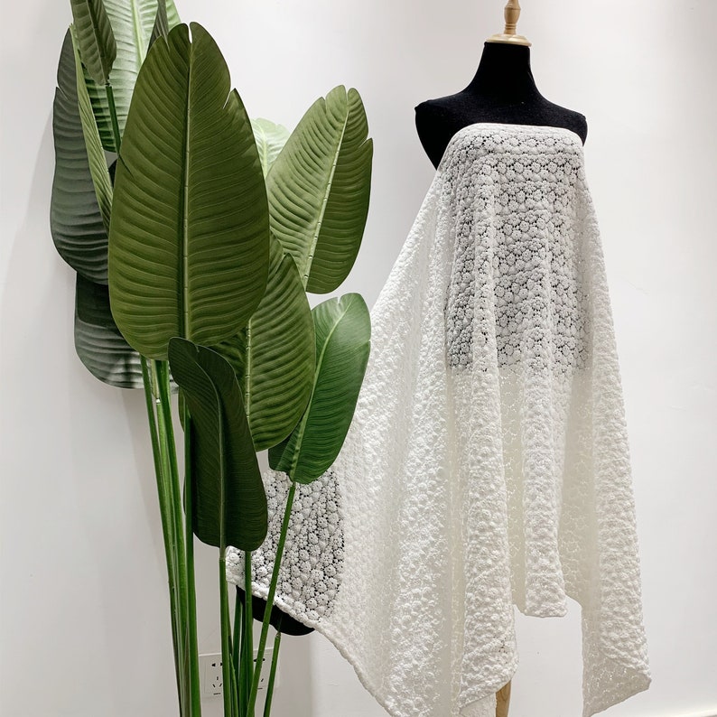 May include: White lace fabric with a delicate floral pattern. The fabric is draped over a mannequin and has a flowing, asymmetrical shape.