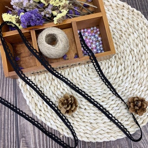 May include: Black elastic trim with a scalloped edge. The trim is laying on a woven straw mat and a wooden box with beads and twine.