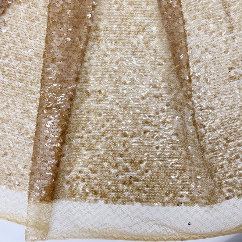 May include: Close-up of a gold sequined fabric with a delicate lace trim. The sequins are small and evenly spaced, creating a shimmering effect.