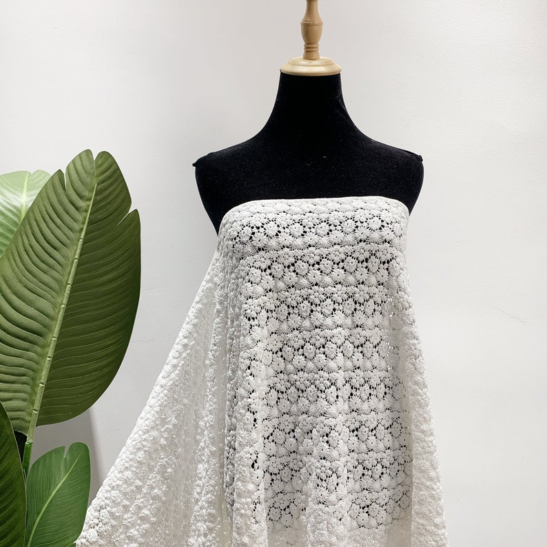 May include: White crochet fabric with a delicate floral pattern. The fabric is draped over a black dress form.