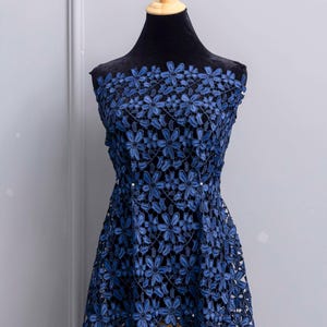 May include: A sleeveless dress featuring a dark blue floral lace overlay with a repeating pattern of six-petaled flowers. The lace is layered over a solid black fabric base, creating a high-contrast look with a defined waistline and a flared skirt.