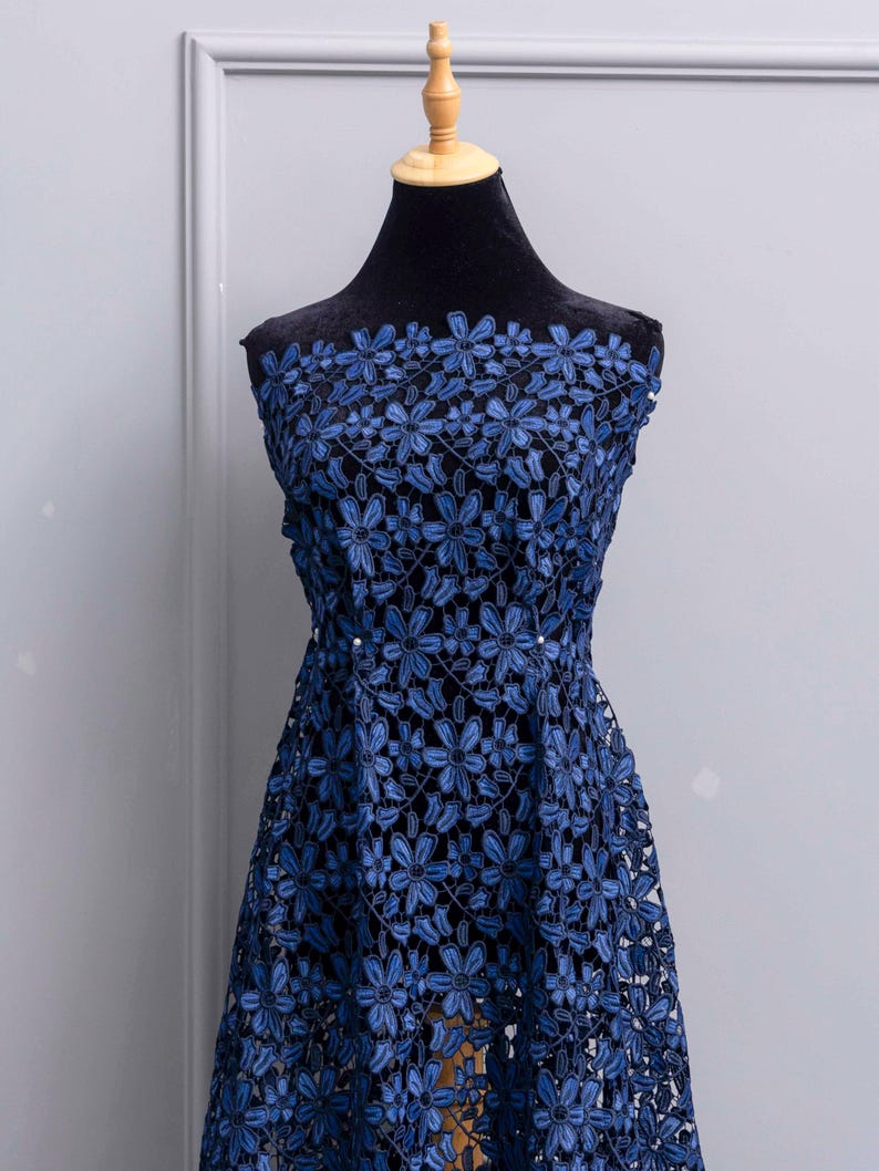 May include: A sleeveless dress featuring a dark blue floral lace overlay with a repeating pattern of six-petaled flowers. The lace is layered over a solid black fabric base, creating a high-contrast look with a defined waistline and a flared skirt.