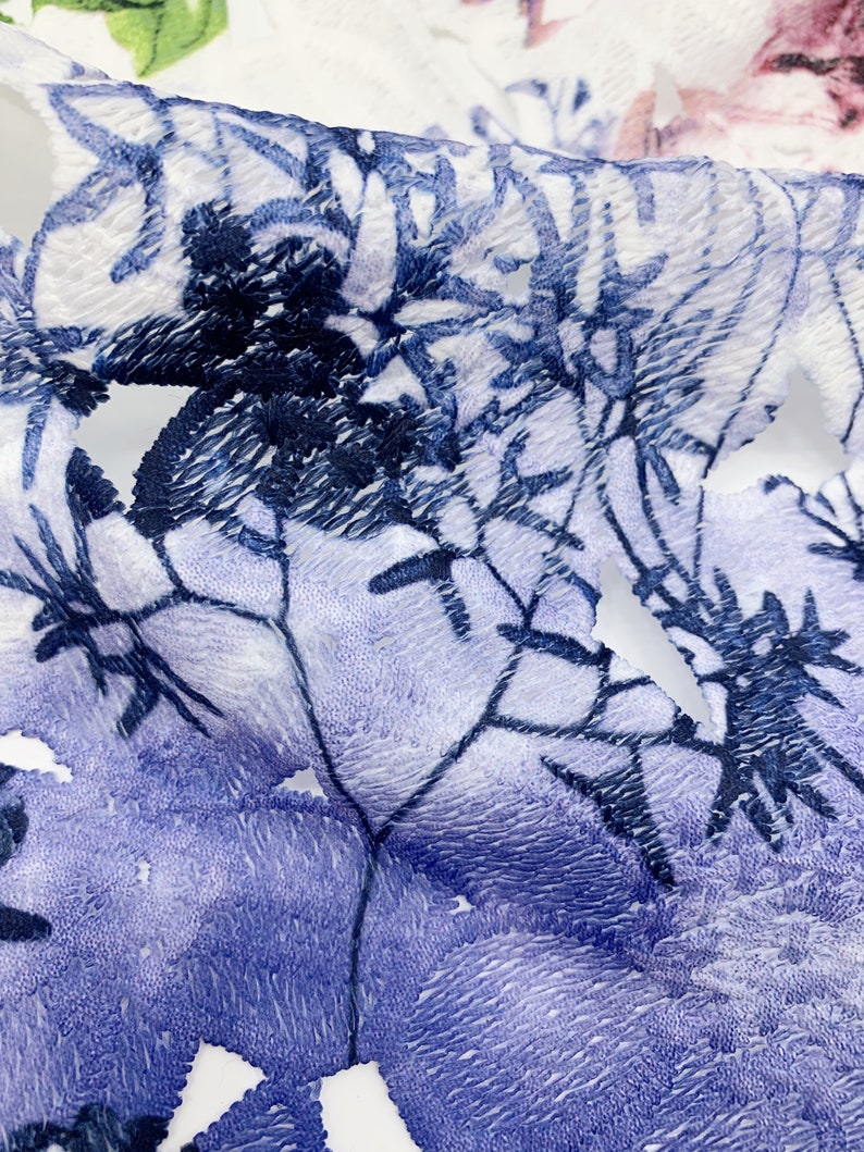 以下が含まれることがあります： 青い刺繍の花柄が施された白い布のクローズアップ。布は質感があり、少し毛羽立った表面をしています。