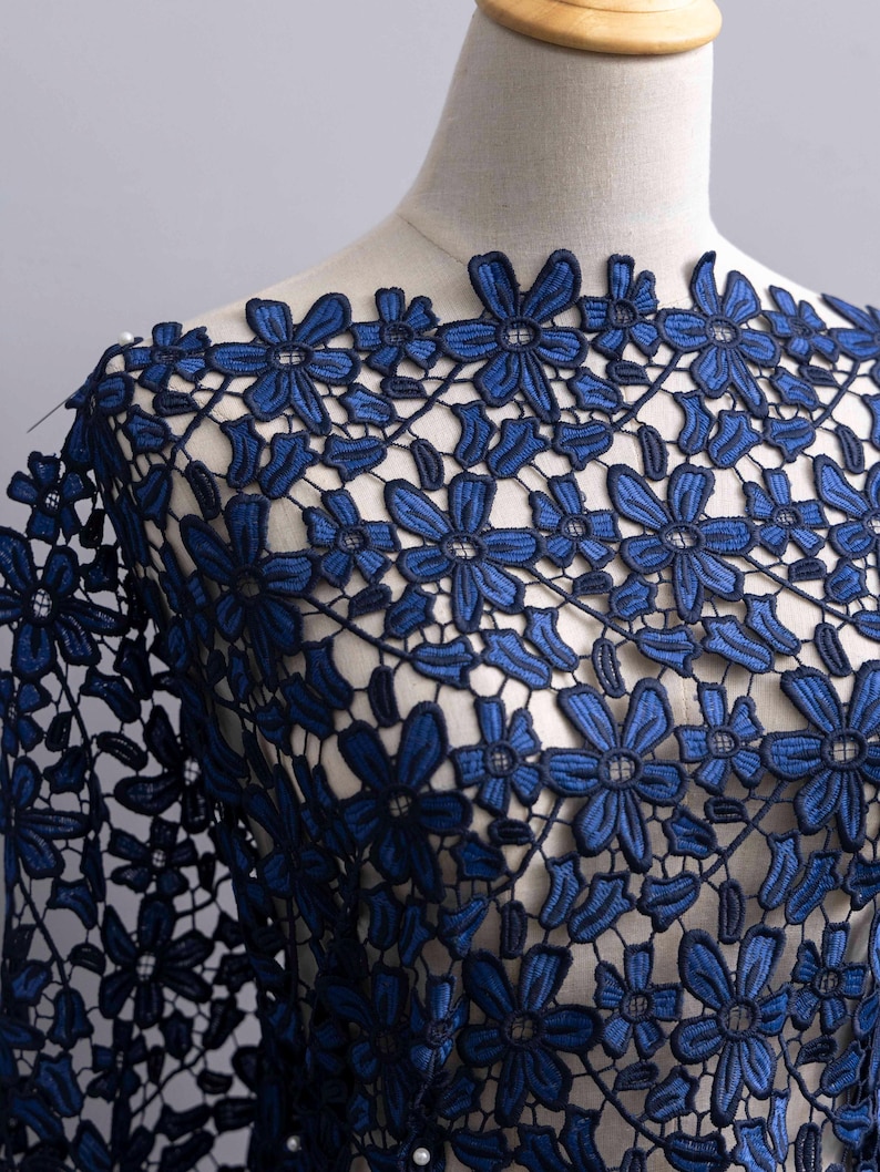May include: A dark blue lace fabric featuring an intricate floral pattern with interconnected petals and stems. The textile is displayed on a mannequin, showcasing the openwork design and the deep navy blue thread used throughout the embroidery.