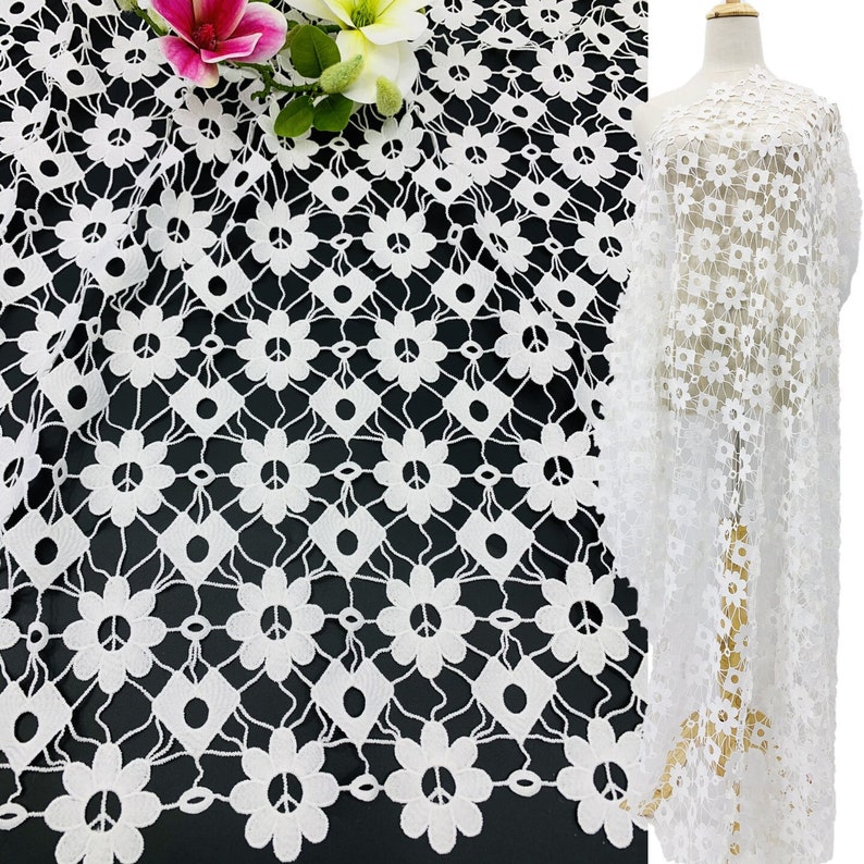 May include: White lace fabric with a repeating pattern of peace signs and flowers. The fabric is shown draped over a surface and on a dress form.